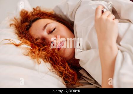 Primo piano di una ragazza rossa che si trova nel letto e dorme. Una ragazza sdraiata su un soffice cuscino coperto da una coperta e dorme stretto. Foto Stock