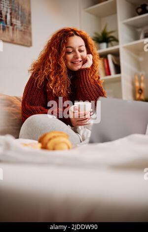 Una ragazza zenzero si siede sul letto al mattino, bevendo il caffè del mattino e usando il suo computer portatile. Foto Stock