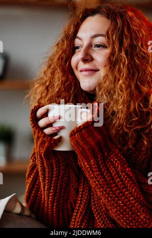 Una giovane donna carina con capelli ricci rossi sta godendo la sua tazza di caffè al mattino. Foto Stock