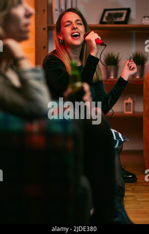 Una giovane donna tiene un microfono e canta una canzone al karaoke party con la sua amica nel soggiorno. Foto Stock