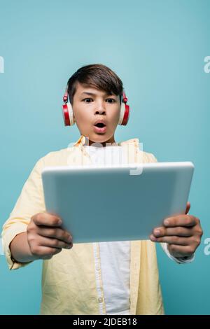 Vista ad angolo basso della pupilla asiatica scioccata in cuffie che tengono un tablet digitale sfocato isolato su blu Foto Stock
