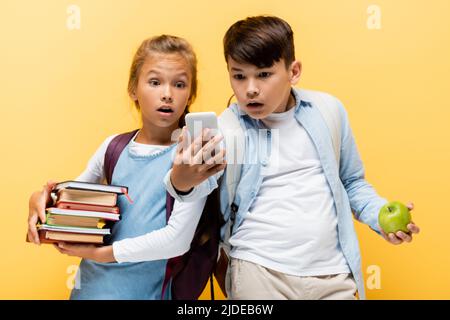 Scioccato scuola interracial bambini con mela e libri guardando smartphone isolato su giallo Foto Stock