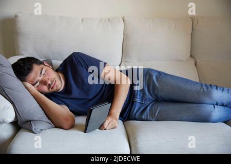 Giovane uomo che si trova addormentato leggendo in un ebook sul divano Foto Stock