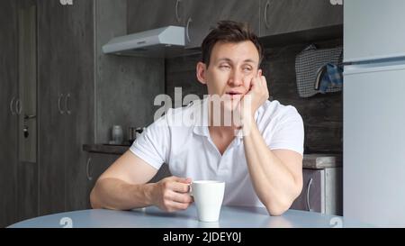 L'uomo assonnato si assapiglia al tavolo e beve una tazza di caffè in cucina Foto Stock