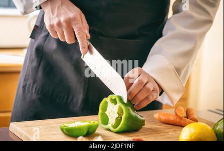 Cuoco con grembiule nero che taglia il pepe verde su un tagliere, cuoci la mano tenendo un coltello vicino. Verdure fresche sul tavolo da cucina, verdure del ristorante Foto Stock