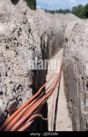 Un fascio di cavi in fibra ottica arancione si trova in una trincea scavata nel terreno in una strada. Mettere a fuoco i cavi in basso a sinistra, sfondo sfocato della Th Foto Stock