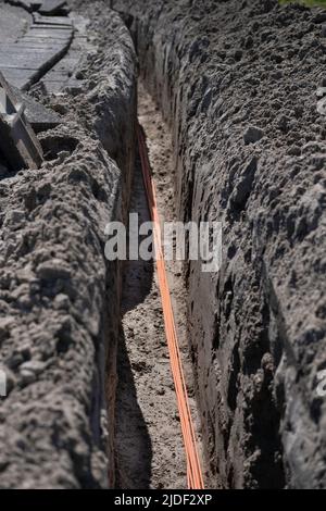 Un fascio di cavi in fibra ottica arancione si trova in una trincea scavata nel terreno di una strada. Internet veloce. Mettere a fuoco i cavi nella parte inferiore della foto Foto Stock