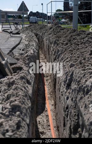 Un fascio di cavi in fibra ottica arancione si trova in una trincea scavata nel terreno di una strada. Internet veloce. Mettere a fuoco i cavi nella parte inferiore della foto Foto Stock