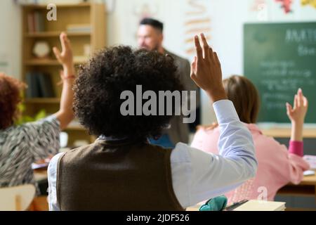 Vista posteriore del ragazzo adolescente afroamericano che alza la mano per rispondere alla domanda dell'insegnante mentre si siede di fronte ai suoi compagni di classe Foto Stock