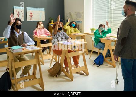 Gli studenti interrazziali in maschere protettive alzano le mani mentre si siedono da scrivanie davanti al loro insegnante in piedi da lavagna a lezione Foto Stock