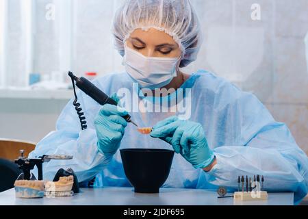 Un dentista in indumenti protettivi sta lavorando su un dente protesico nel suo laboratorio. Foto Stock