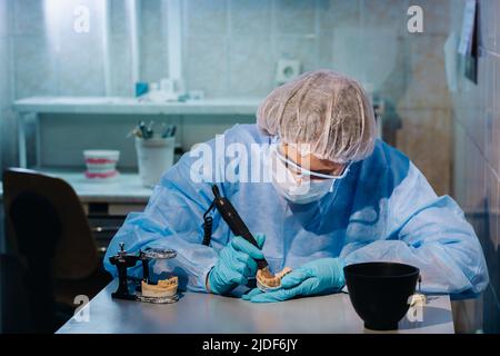 Un dentista in indumenti protettivi sta lavorando su un dente protesico nel suo laboratorio. Foto Stock