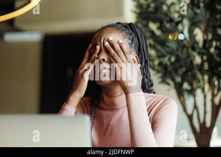 Giovane donna afro americana infelice con paurini che piangono gli occhi coprendo con le mani in chioseup caffè casa. Depressa ragazza nera che soffre, disperata Foto Stock