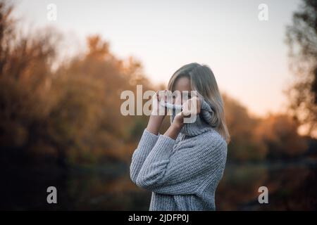 Donna bionda affascinante in piedi dal fiume nella foresta, guardando via sorridente e tirando il collare maglione sopra faccia con gli alberi coperti con il tramonto dentro Foto Stock