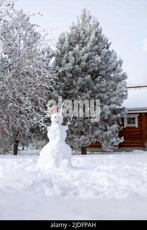 Uomo di neve fatto a mano fuori su terreno coperto di neve con pini e casa di legno di campagna in background in giorno. Giocare all'aperto in inverno Foto Stock