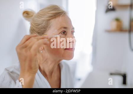 Bella donna anziana in accappatoio, pelle idratante con pipetta con trattamento siero godendo procedura. Foto Stock