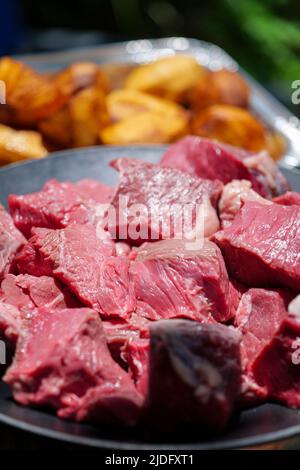 Primo piano di pezzi di carne di manzo crudo a dadini su un piatto di fogli e patate arrosto sullo sfondo. Foto Stock