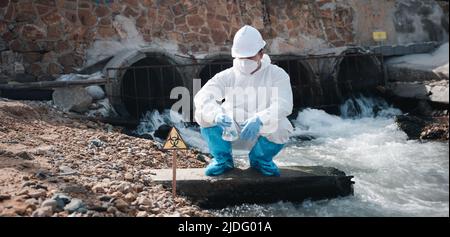 Ecologo campionamento acqua sostanze chimiche tossiche dal fiume con vetro provetta e hanno fumo bianco, Biologo indossare tuta protettiva e maschera raccoglie il campione Foto Stock