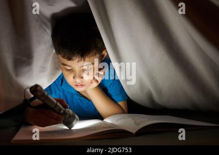 Ragazzo preteen che usa la torcia quando legge il libro sotto coperta a tarda notte Foto Stock