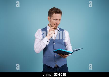 Ritratto positivo bello giovane assistente di affari con una barba in piedi su uno sfondo blu in vestiti di affari e fare note in un diario Foto Stock