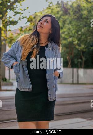 Una donna latinoamericana dai capelli lunghi che indossa una giacca jean ha le mani nelle sue tasche e guarda gli alberi alla stazione del tram Foto Stock