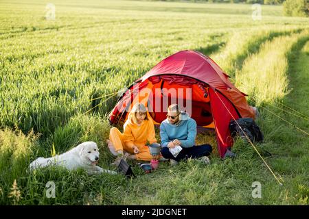 La giovane coppia ha un pic-nic con un cane vicino alla tenda sul campo verde Foto Stock