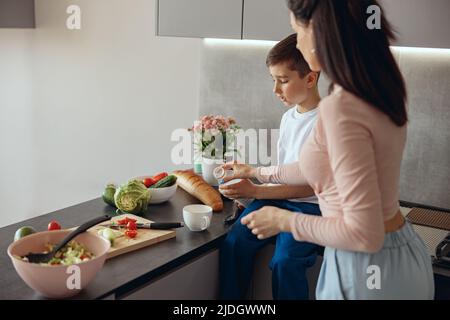 Dietro la madre caucasica che insegnava al ragazzino a cucinare. Figlio con mamma che versa il latte in una tazza. Foto Stock