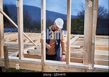 Uomo lavoratore costruire casa di legno telaio su palo fondazione. Il carpentiere martella il chiodo in una travetta di legno, usando il martello. Concetto di carpenteria. Foto Stock