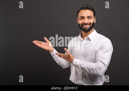 L'imprenditore indiano maschio punta con entrambe le mani a parte isolato sul nero, uomo d'affari bearded che presenta, mostrando la novità guardando la macchina fotografica e sorrisi, spazio copia Foto Stock