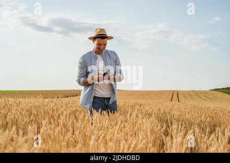 Un agricoltore felice che usa il telefono cellulare mentre si trova nel suo campo di grano in crescita. Foto Stock