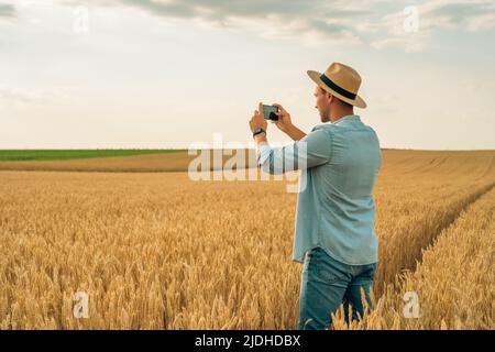 Felice agricoltore fotografando le colture con il telefono mentre si trova in piedi nel suo campo di grano crescente. Foto Stock