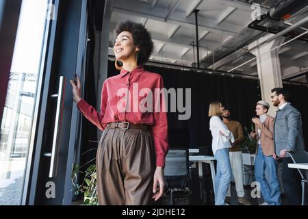 vista ad angolo basso dell'agente pubblicitario afro-americano positivo che guarda via vicino ai colleghi interrazziali in ufficio Foto Stock