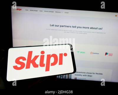 Persona che tiene uno smartphone con il logo della società belga di mobilità Skipr SA sullo schermo di fronte al sito web. Mettere a fuoco sul display del telefono. Foto Stock