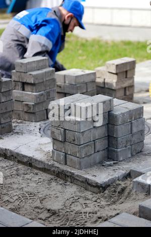 Il muratore in abiti da lavoro si siede sul marciapiede e stende le lastre di pavimentazione. Vista dell'uomo in attività all'aria aperta. Costruttore professionista fa organizzazione del territorio giorno d'estate. Scena reale. Foto Stock