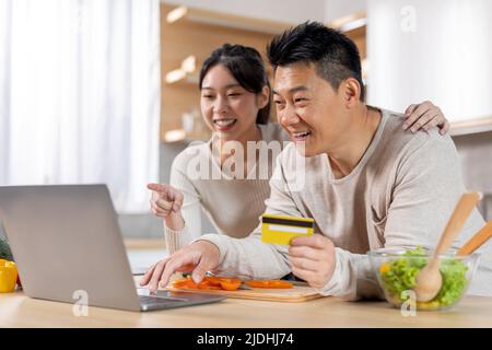 Coppia asiatica eccitata che fa l'ordine in linea, usando il laptop alla cucina Foto Stock