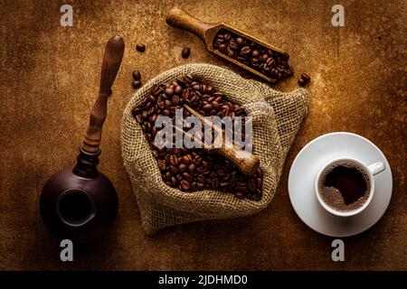 Composizione del caffè a disposizione piatta. Sacco di burlap con chicchi di caffè organico torrefatto, bevanda americana appena fatta in tazza, ciotola di cezve, cucchiaio di legno sopra la grung Foto Stock