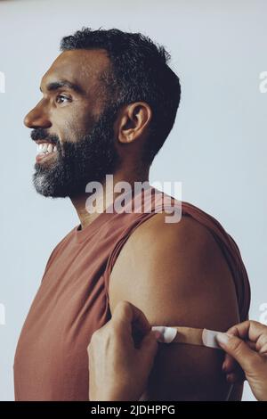 profilo adulto medio sorridente bearded vaccinato con le mani che collocano l'intonaco sul braccio uomo nello studio colpo guardando via Foto Stock
