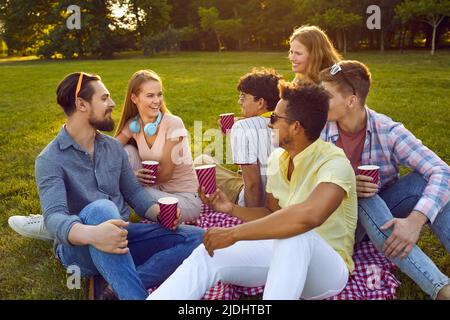 Felici amici multietnici seduti sul prato verde con coppe di carta nella giornata estiva soleggiata. Foto Stock