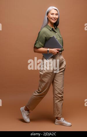 Donna sorridente positiva con capelli grigi in piedi con un computer portatile chiuso in mano, mentre si è in pausa. Studio interno girato isolato su sfondo beige Foto Stock