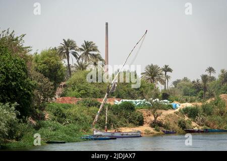 Una feluca tradizionale con diverse piccole barche ormeggiate nelle canne sulle rive del Nilo con muratura e camino sullo sfondo Foto Stock