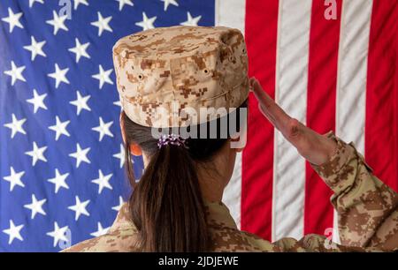 Donna US Army Soldier in piedi di fronte a una bandiera americana e saluta. Donna in uniforme militare vista posteriore Foto Stock