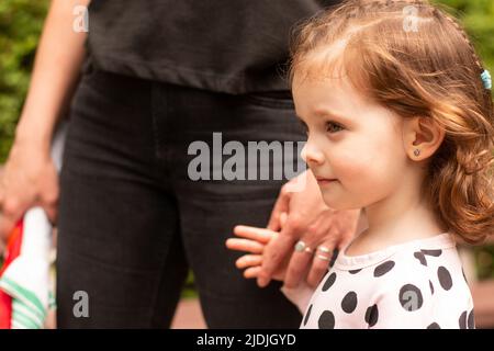 Piccola bella ragazza dai capelli rossi con capelli ricci in un abito tiene la mano di un genitore in un parco estivo Foto Stock