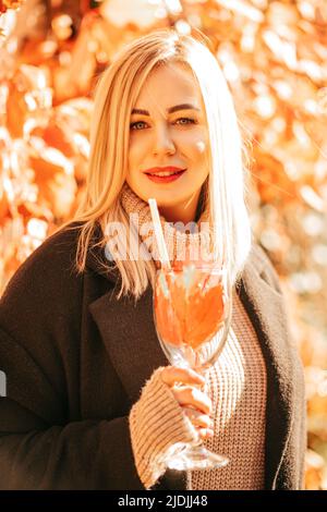 La lussuosa bionda in cappotto nero e maglione nel parco festeggia l'arrivo dell'autunno con un bicchiere di cocktail autunnale fatto di foglie luminose Foto Stock