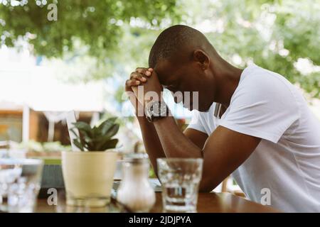 Verticale stressato uomo pelle scura appoggiando la testa su mani congiunte vicino al laptop in caffetteria. Mal di testa ed emicrania perché lavoro Foto Stock