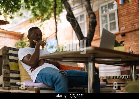 Serio, concentrato ed entusiasta afro americano uomo usando il laptop e bere caffè in internet cafe. Fai affari Foto Stock