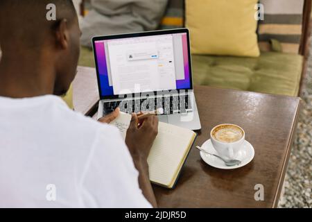 Uomo interrazziale retroguarista che utilizza un computer portatile, scrive note in un libro di testo e beve caffè in un caffè con tecnologia wireless. Foto Stock