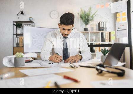 Competente arhitect indiano maschio seduto sul tavolo interno utilizzando penna e righello per i disegni di progetto. Giovane uomo indù Positiv con camicia bianca e cravatta nera. Foto Stock