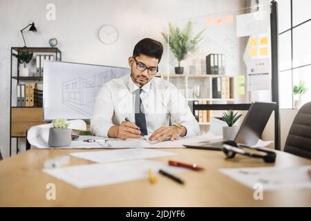 Architetto arabo qualificato in maglia bianca, cravatta e occhiali di controllo progetto di costruzione con Blueprint e laptop seduto sul tavolo. Un attraente uomo musulmano che controlla il processo di lavoro. Foto Stock