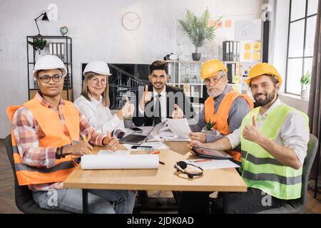 Architetti, ingegneri e designer qualificati che si incontrano in ufficio davanti allo schermo TV. Colleghi multiculturali in abiti e caschi seduti al tavolo e guardando la macchina fotografica. Foto Stock