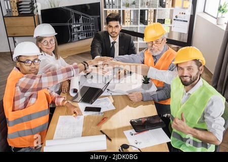 Felici architetti, ingegneri e designer multirazziali che si accatastano le mani durante un incontro di successo nella sala riunioni. Colleghi uomini e donne che conducono il meeting per il progetto architettonico. Foto Stock